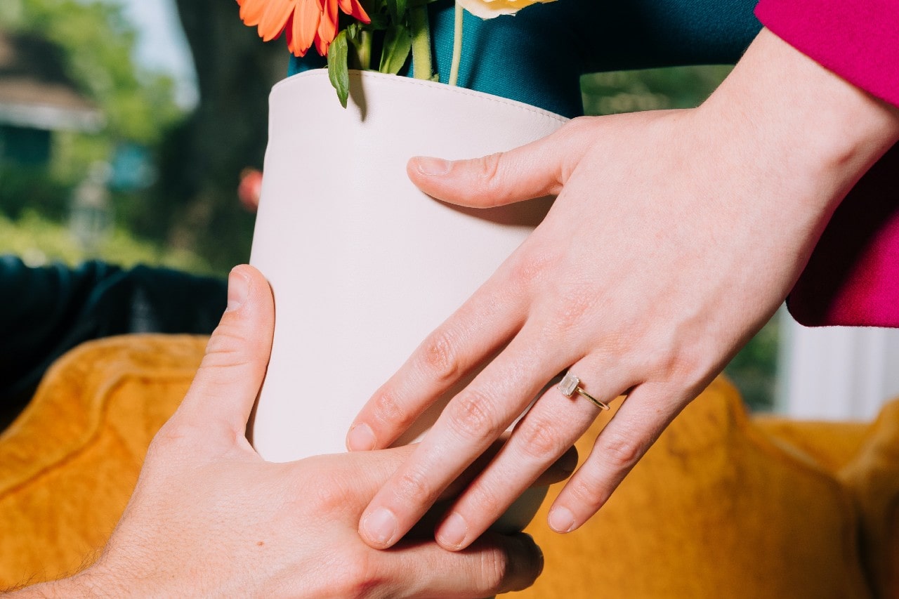 A man passes a flowering plant to his fiancee, wearing an emerald-cut solitaire ring.