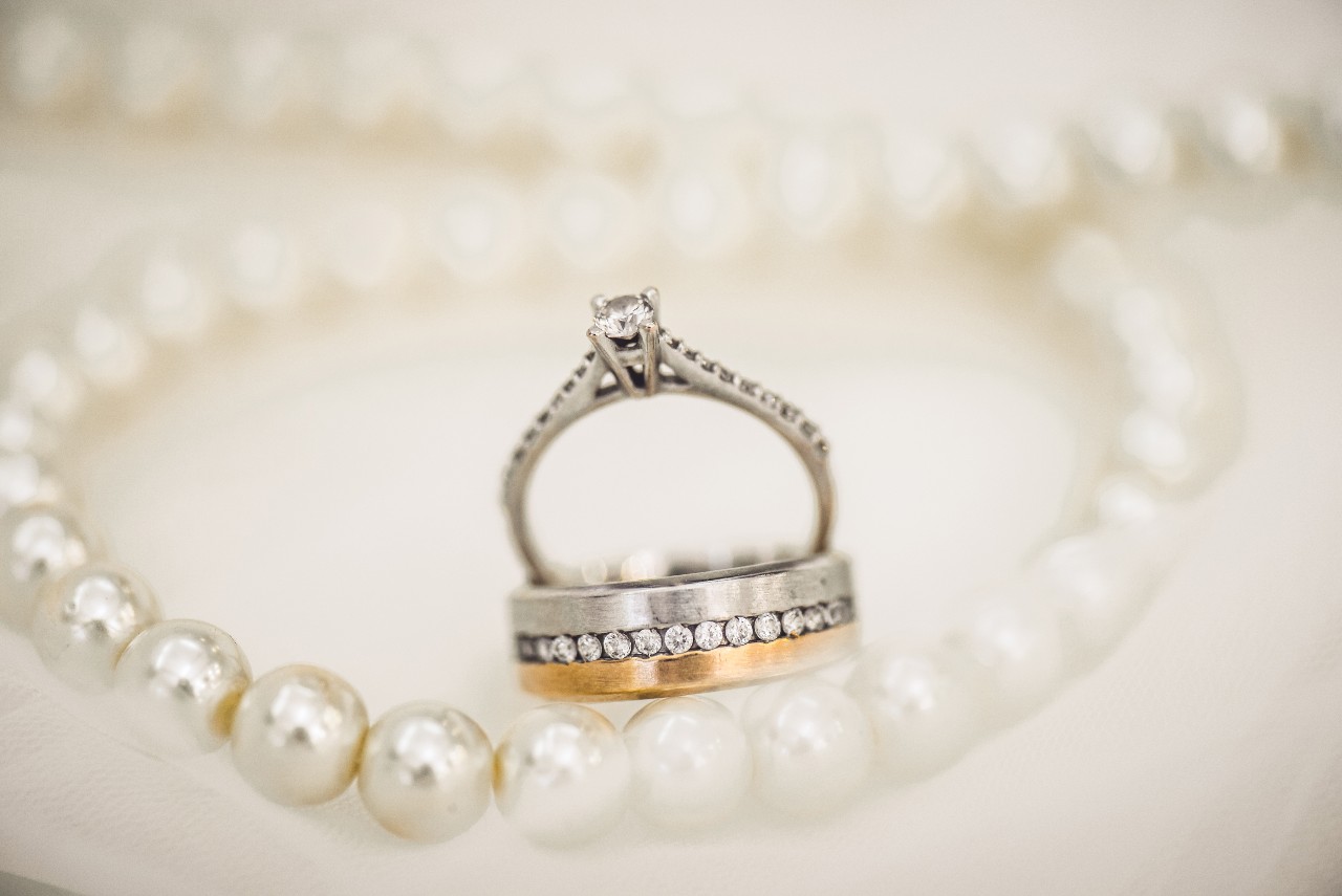 A vintage engagement ring sits inside a mixed metal men’s wedding band and a pearl necklace.
