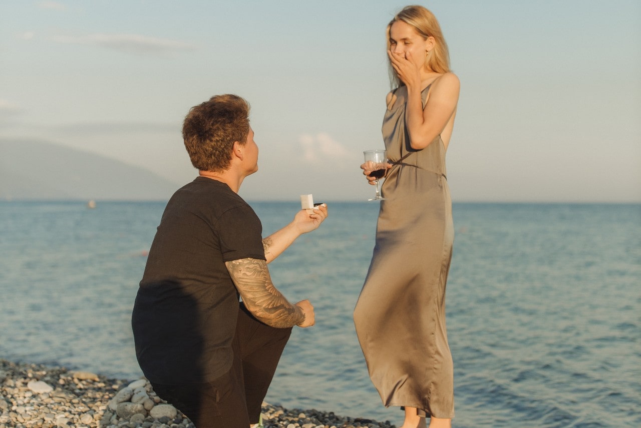 A man proposing to a woman in a long, silk dress holding a glass of wine while she stands on the shore in front of a body of water
