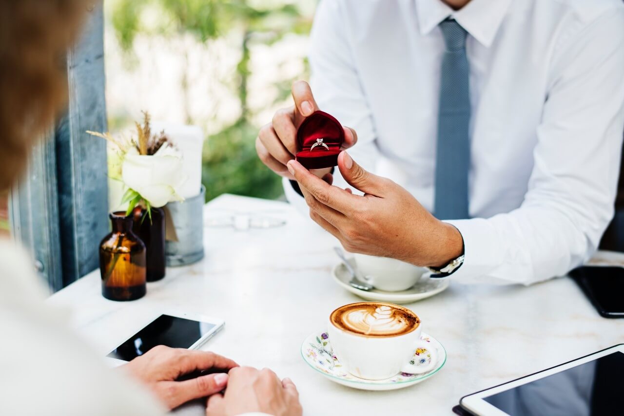 A man proposing with a heart-shaped box with a side stone ring over lattes at a cafe