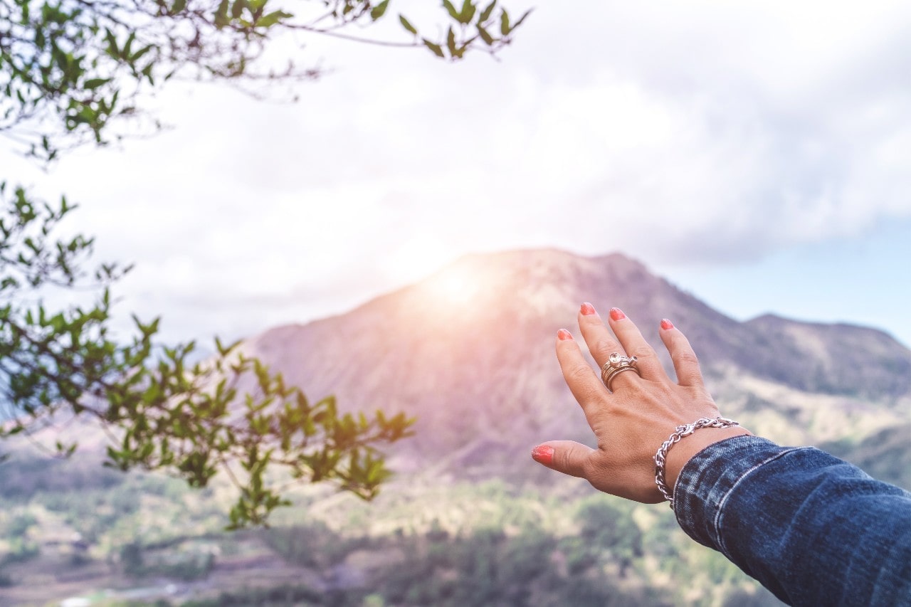 In the foreground, a hand covered in jewelry protects one’s eyes against the sun. A mountain looms in the background