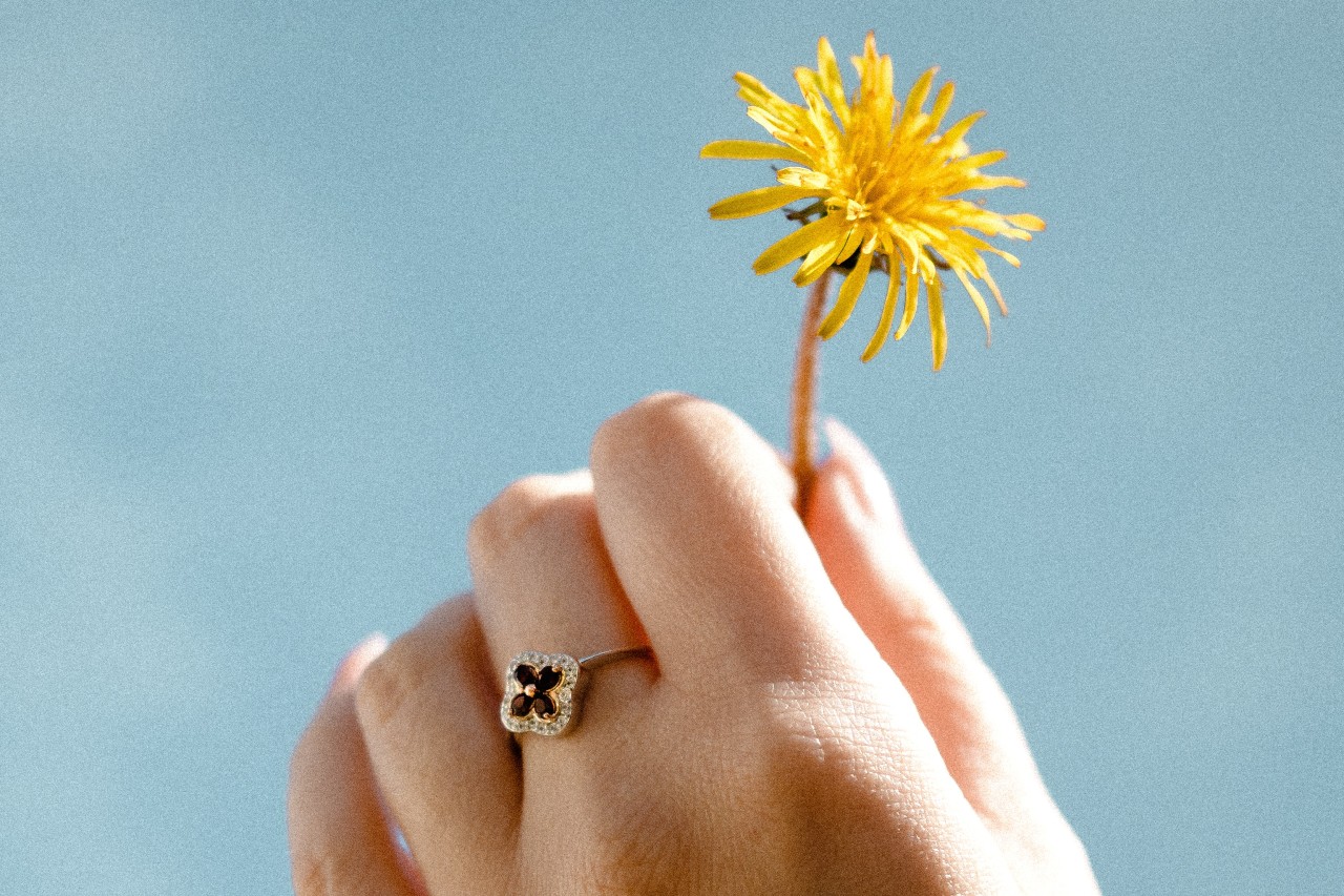 Close up image of a hand holding a dandelion and wearing a floral ring
