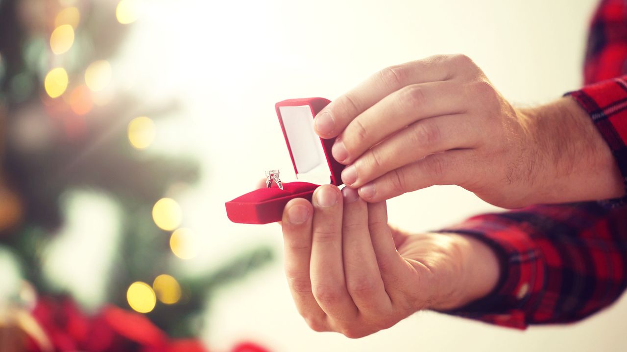 A man in plaid presents an engagement ring in front of a Christmas tree.