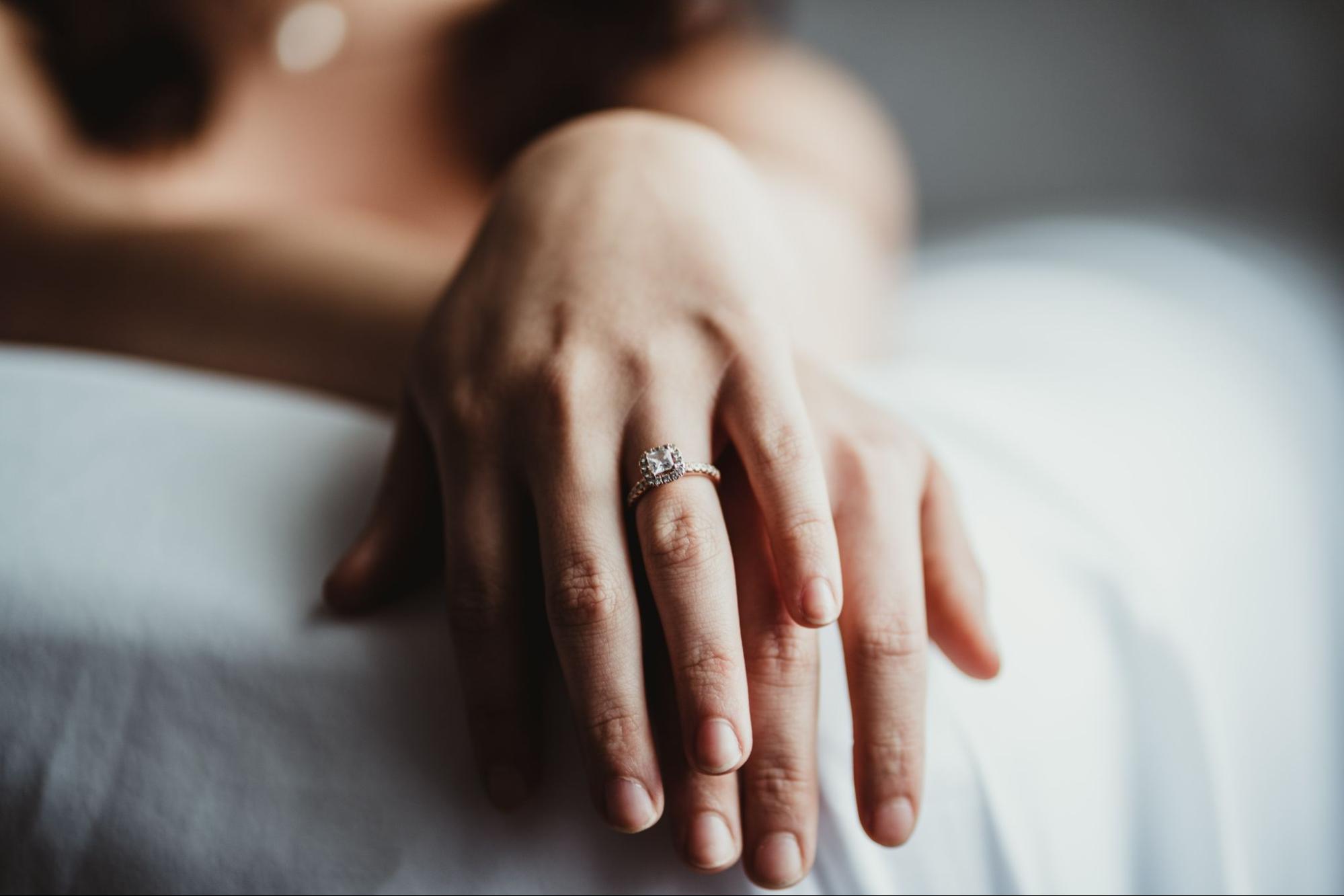 A woman’s hand featuring a princess-cut diamond engagement ring sits on top of a man’s hand.