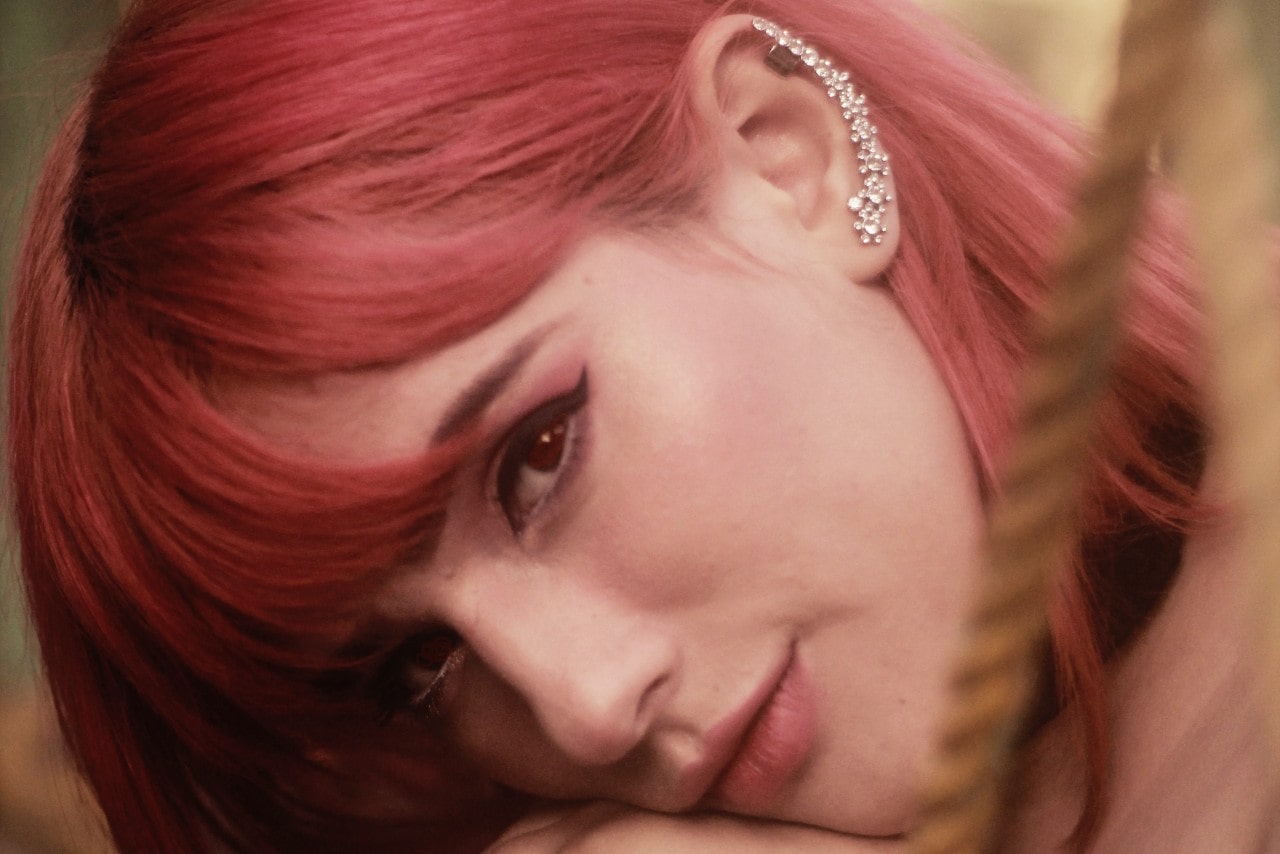 A pink-haired woman wears a diamond crawling earring while resting her head on her arms.